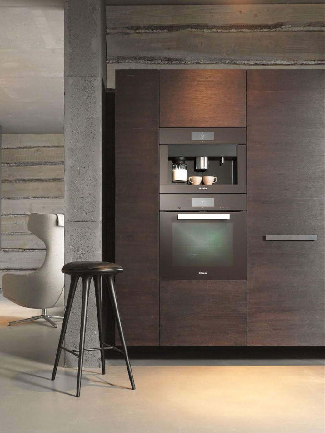 Modern kitchen corner with dark wood cabinetry, built-in coffee machine, oven, black stool, and light concrete floor.