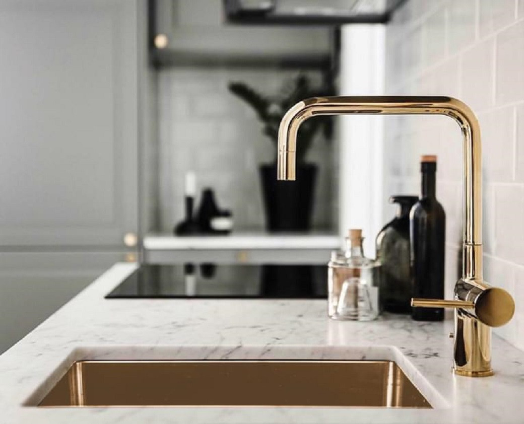 Marble kitchen countertop with gold faucet and sink, black bottles, and blurred background items.