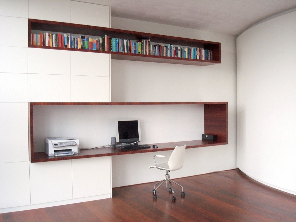 Minimalist home office with dark wood built-in desk and shelves, white cabinets, computer, printer, and white swivel chair.