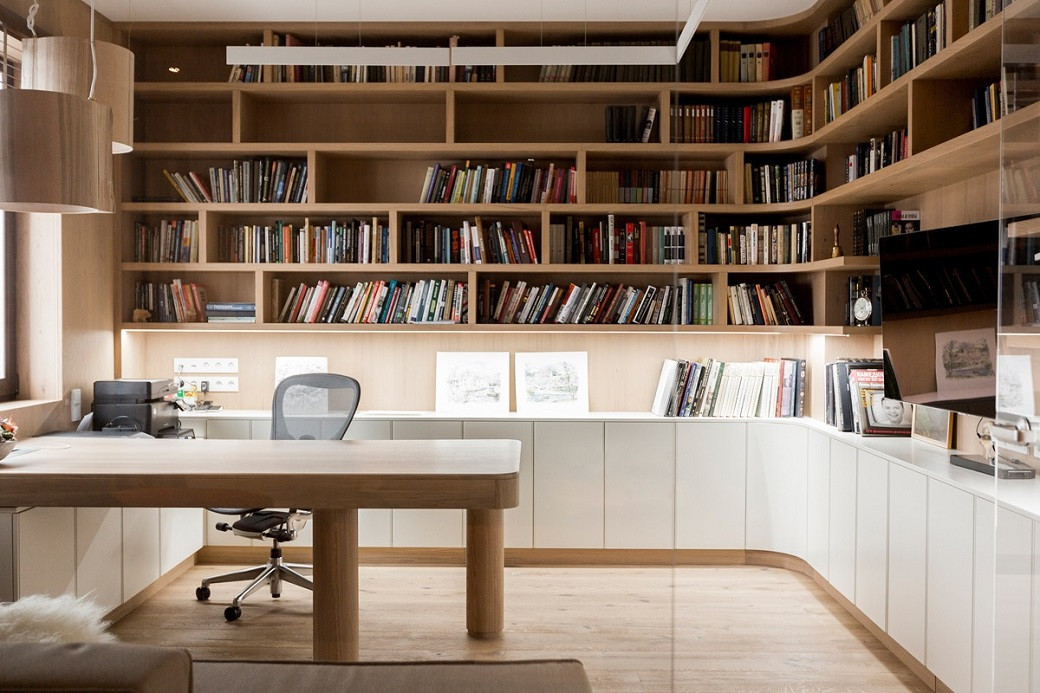 Modern home office with wooden shelves filled with books, a large wooden desk, ergonomic chair, and white cabinets.