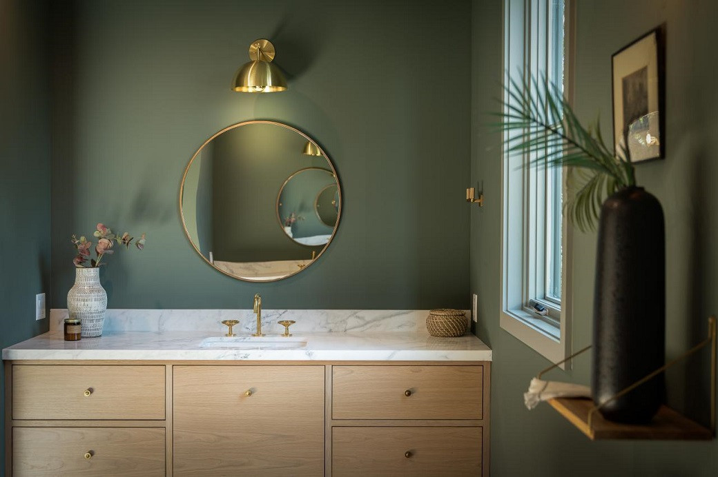 Bathroom vanity with wooden drawers, white marble countertop, gold fixtures, round mirror, green walls, and decorative vases.