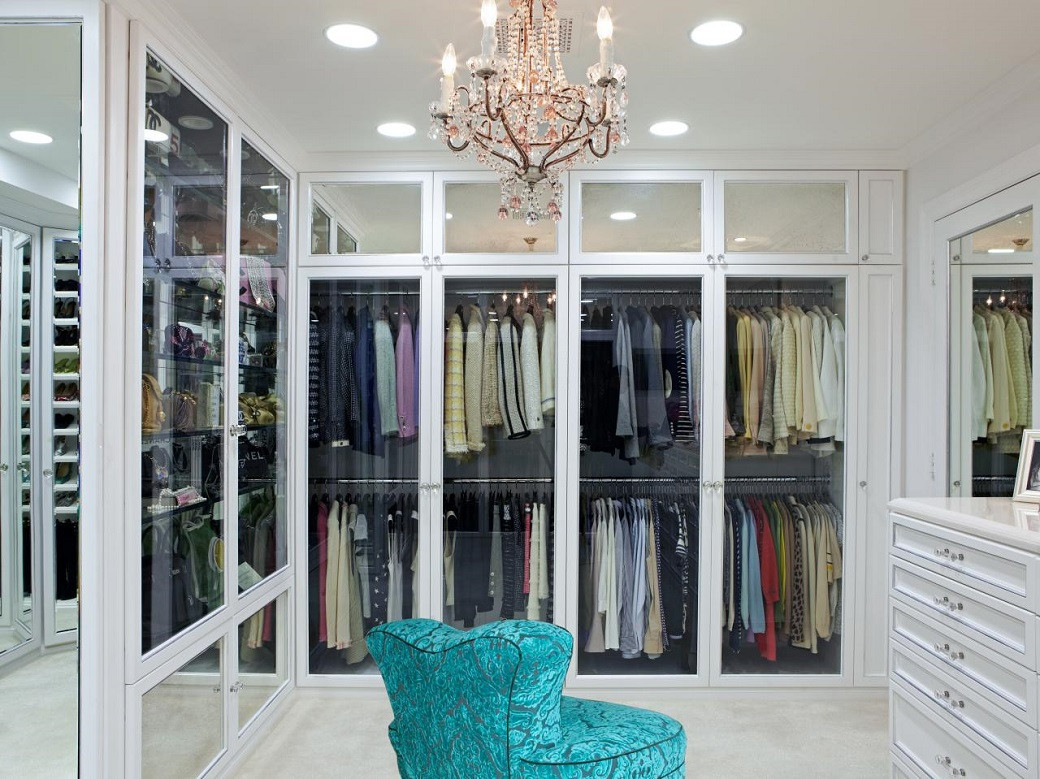 Walk-in closet with white cabinetry, glass doors displaying hanging clothes, turquoise upholstered chair, and chandelier.