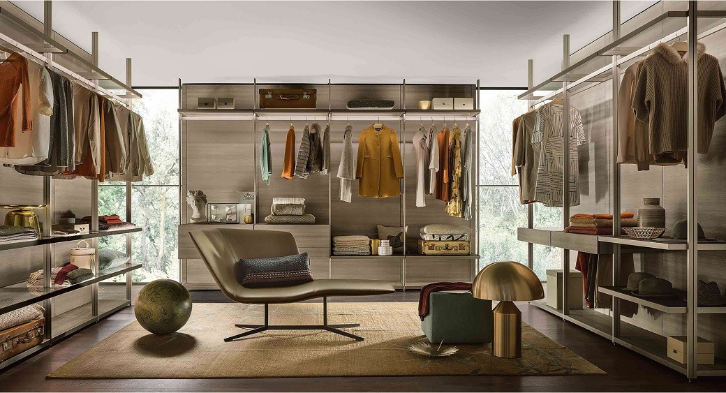 Spacious walk-in closet with open shelving, hanging clothes, folded garments, a lounge chair, a round sculpture, and a gold l