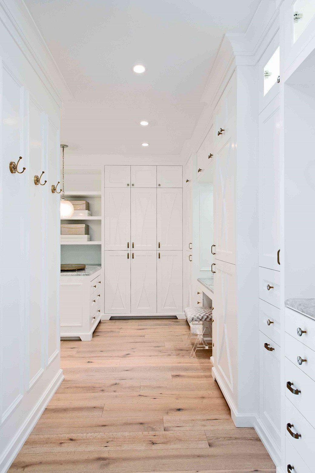 Bright walk-in closet with white cabinetry, brass hooks and handles, wooden floor, shelves, and a small upholstered stool.