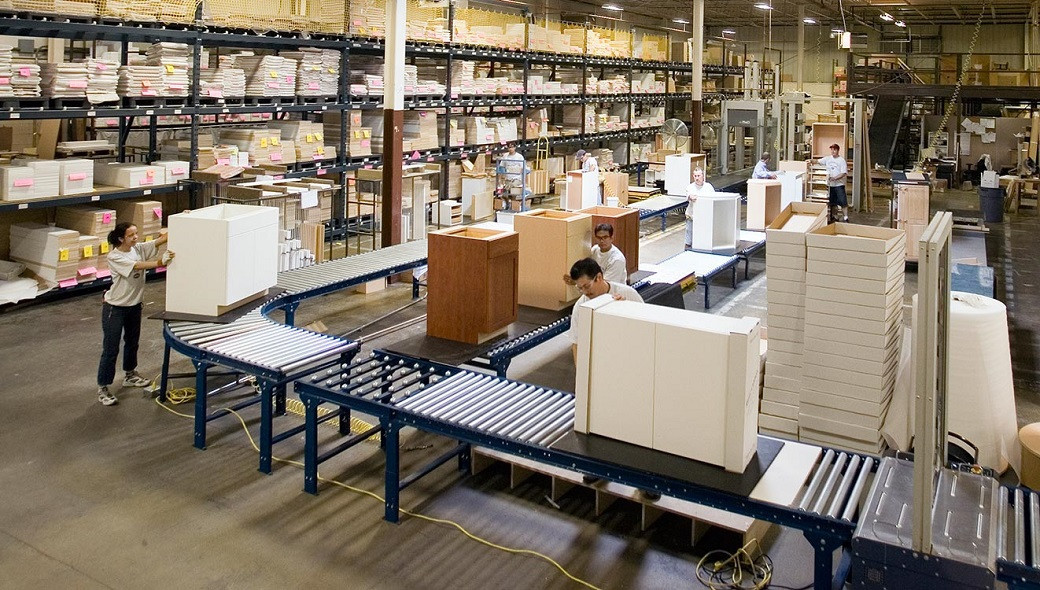 Warehouse interior with workers assembling and handling kitchen cabinet units on roller conveyors.